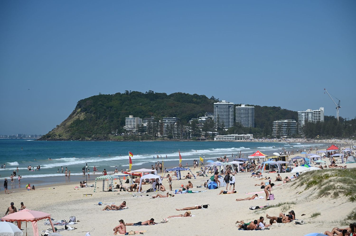 Crowded sandy beach with sunbathers, colorful umbrellas, rolling waves, and a green headland with hotels in the background.