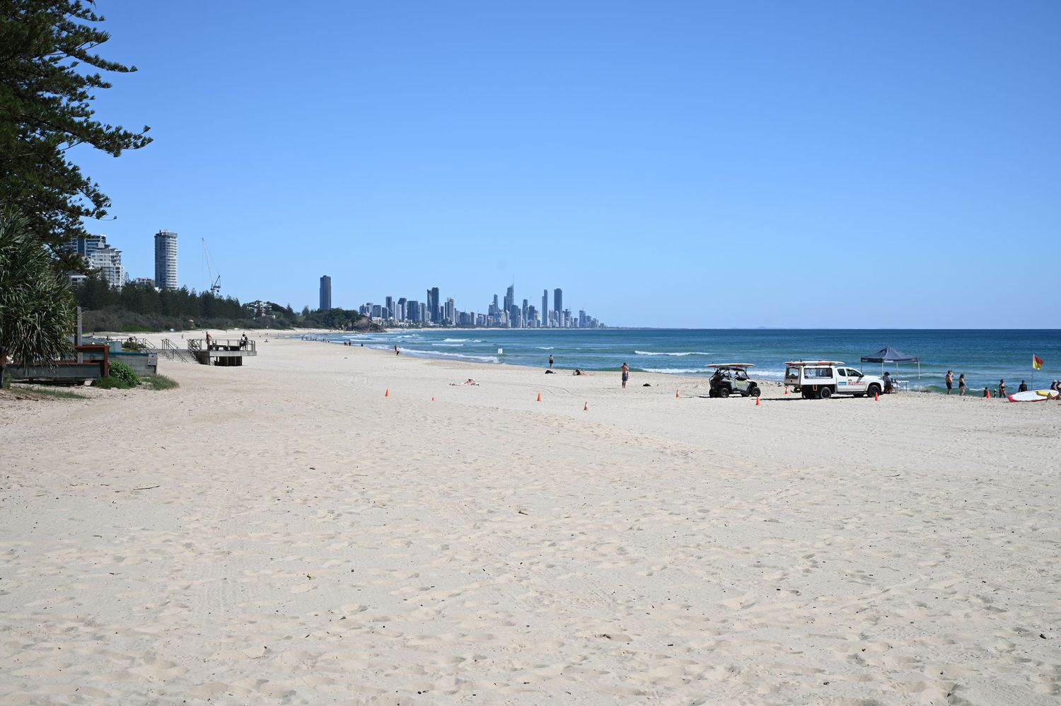 White sandy beach with calm waves, city skyline in background, vehicles on shore, clear blue sky, Gold Coast Australia.