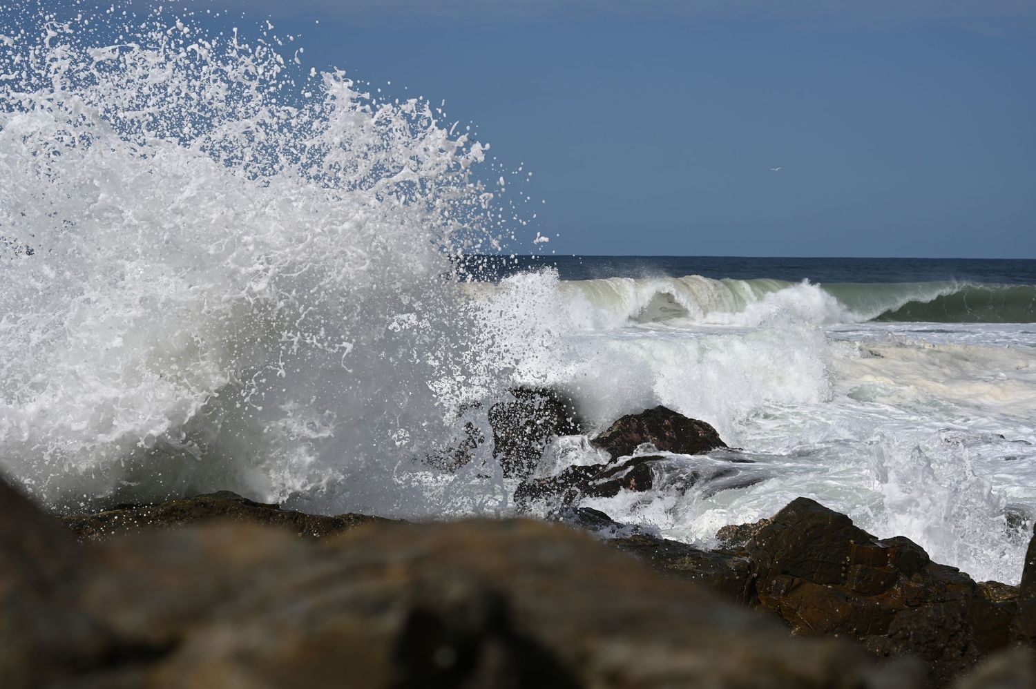 Powerful ocean waves crashing against rocky shoreline with white sea spray under a clear blue sky.