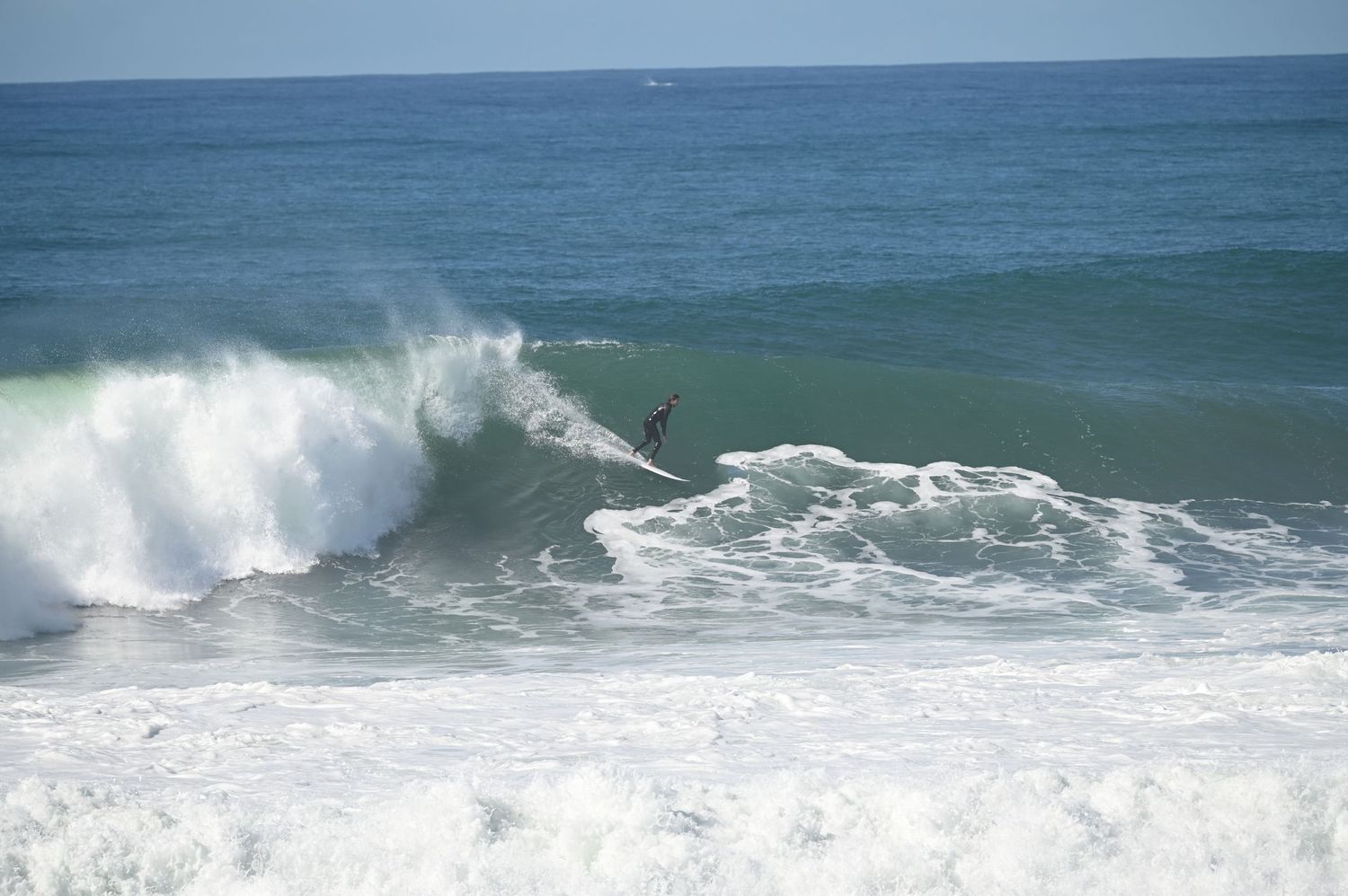 A surfer rides a large crashing wave in the ocean on a sunny day.