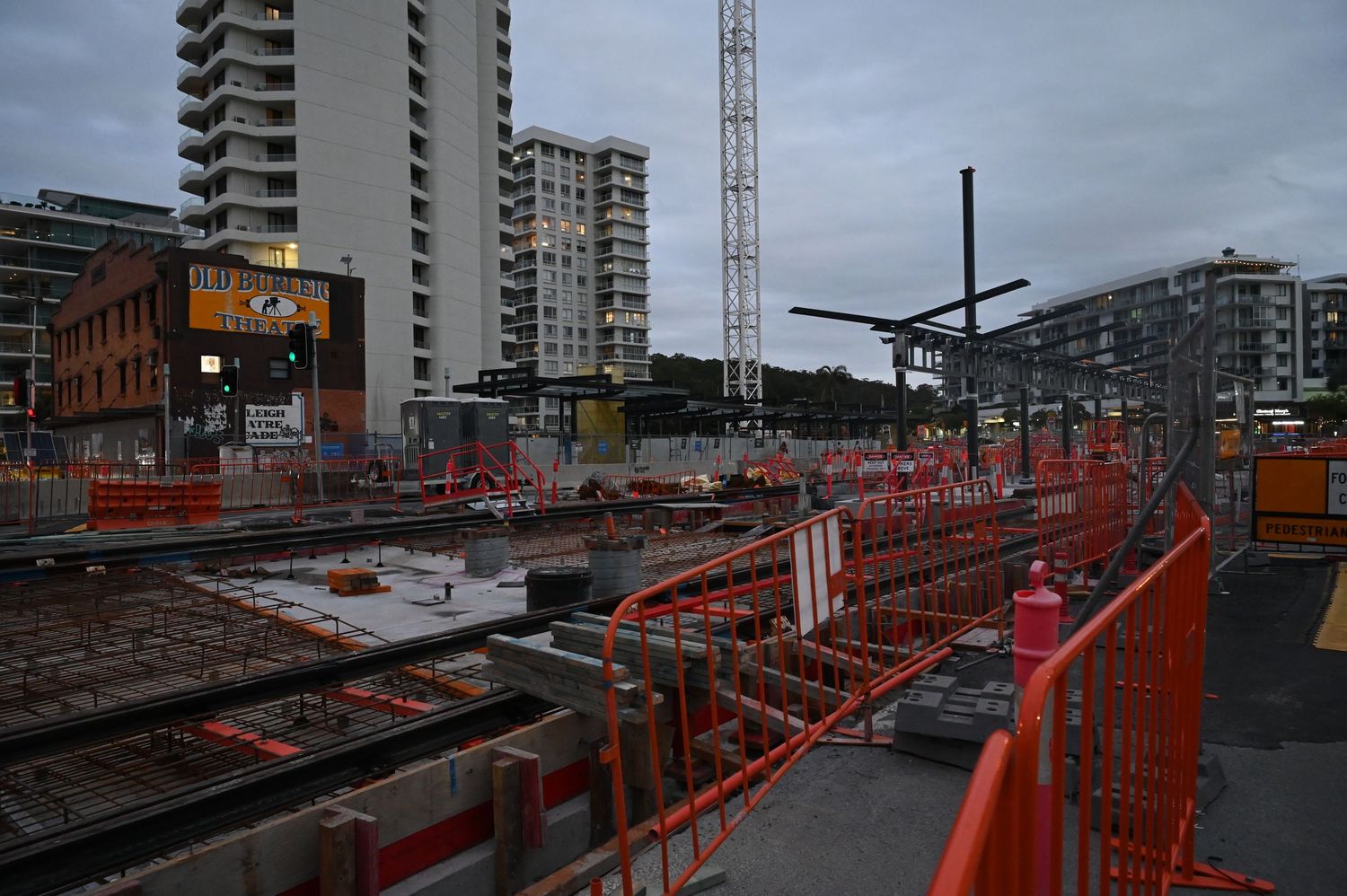 Large urban construction site with orange barriers, cranes, and high-rise buildings at dusk.
