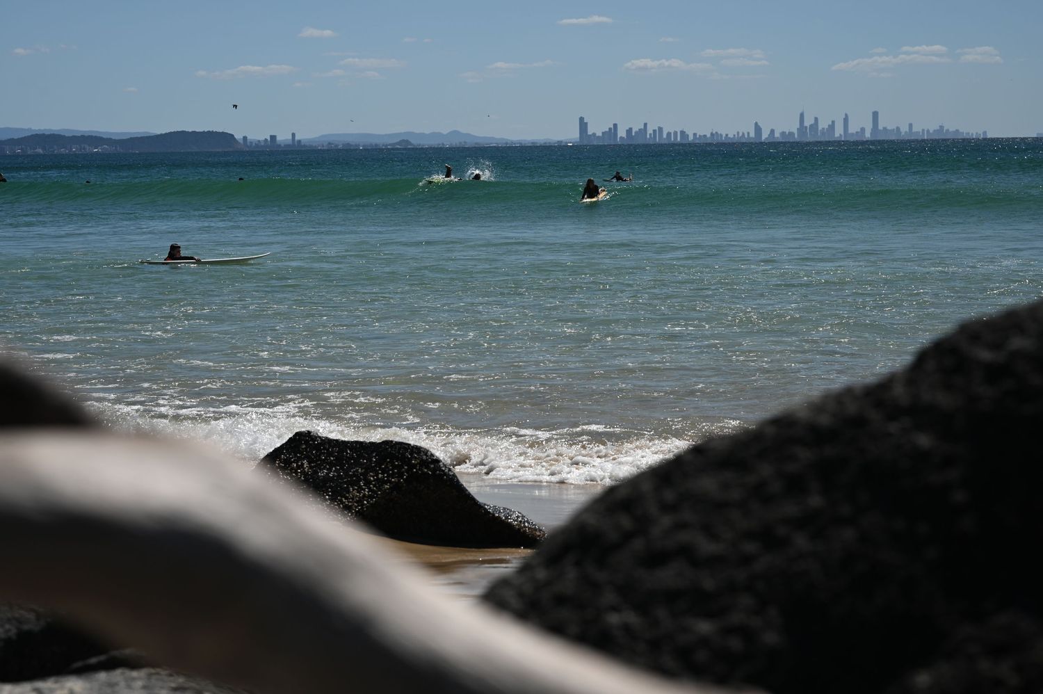 Surfers paddling in calm turquoise ocean waters with a city skyline visible in the distance on a sunny day.
