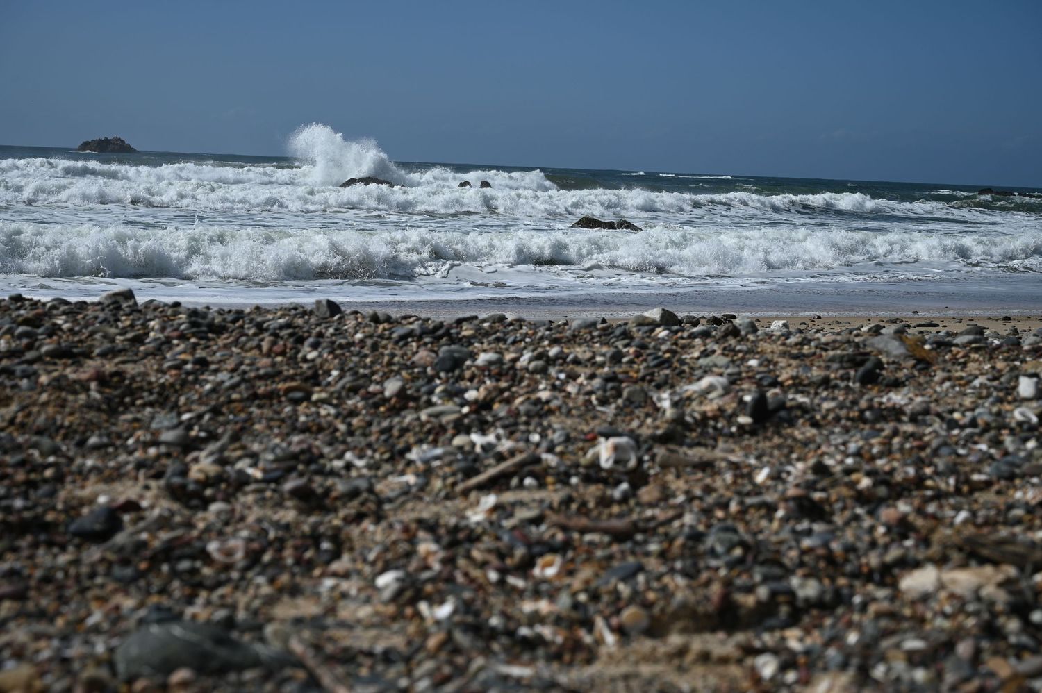 Rocky pebble beach shoreline with crashing ocean waves under a clear blue sky on a sunny day.
