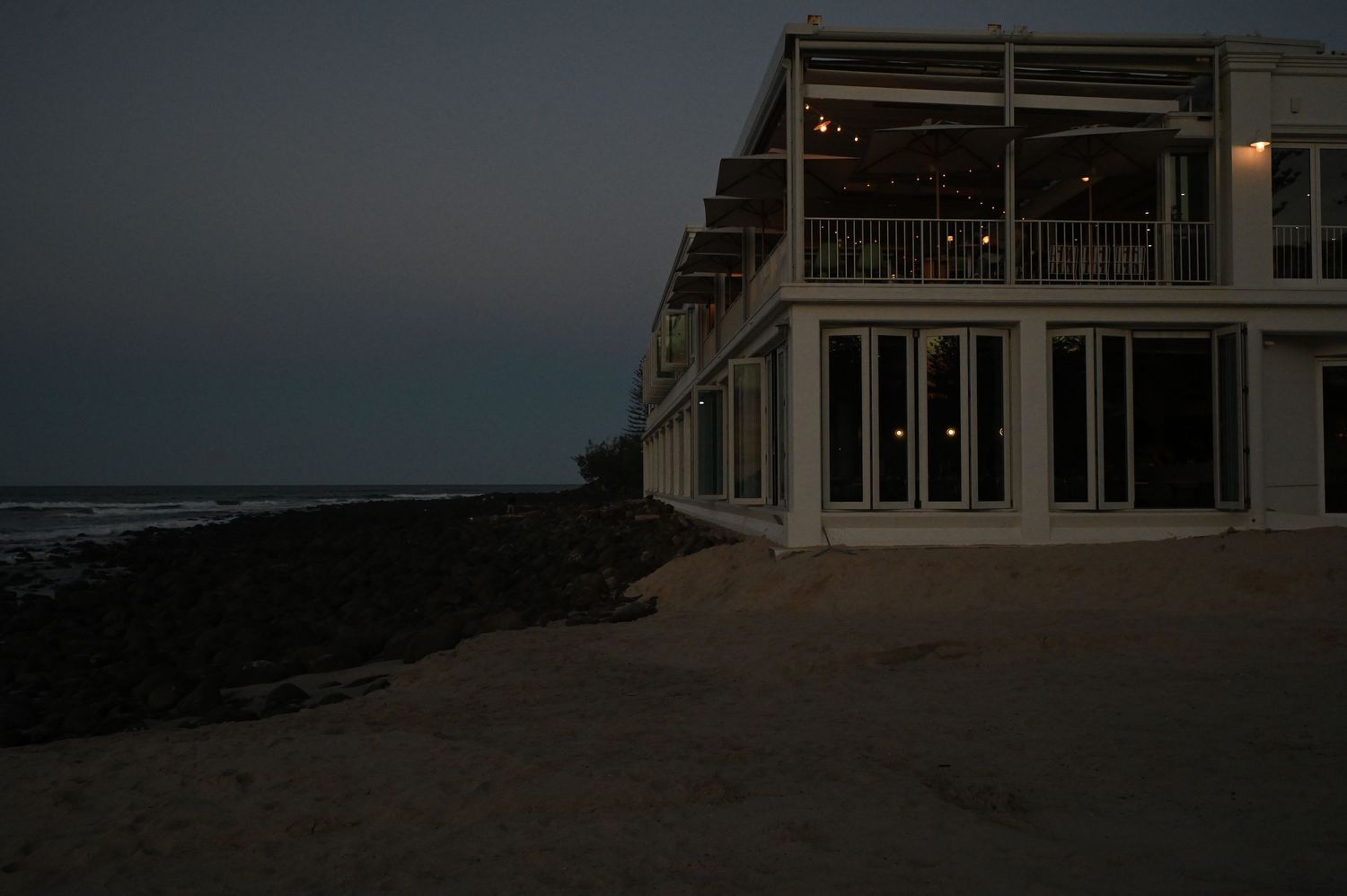 Beachfront restaurant at dusk with lit interior, sandy shore, rocky coastline, and ocean waves in the background.