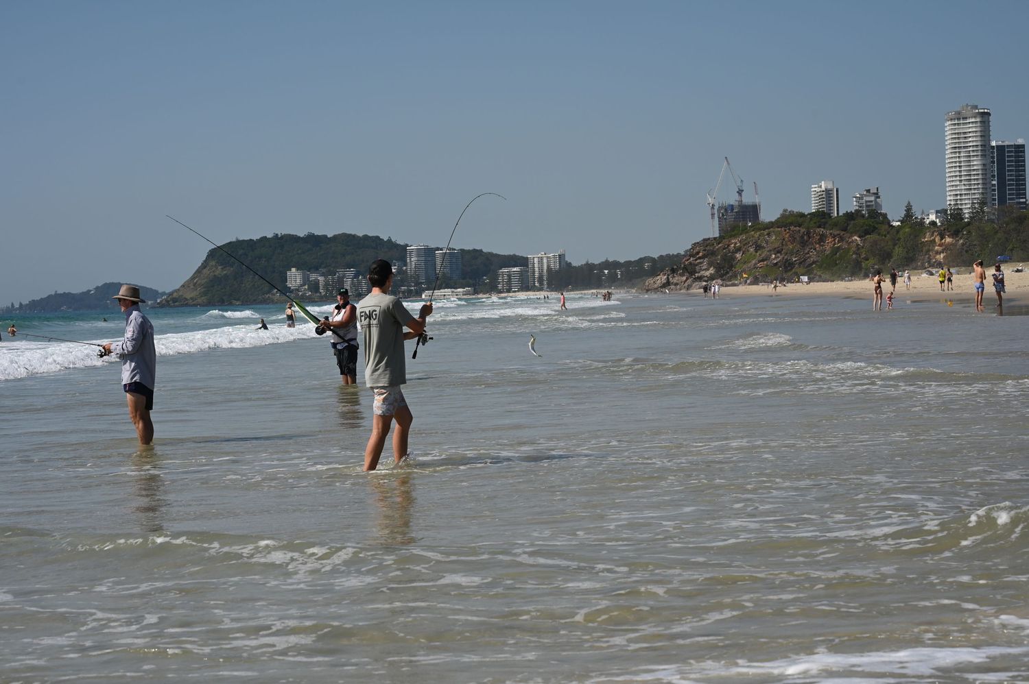 Two people fishing in ocean waves on a sunny beach with city skyline and green headland in background.