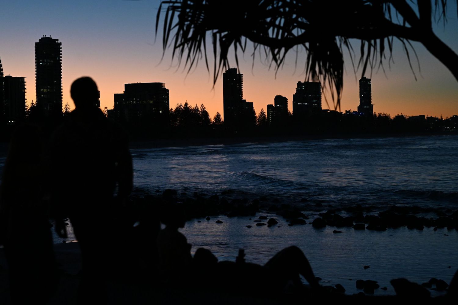 Silhouette of a person standing near rocky shore with city skyline at dusk.