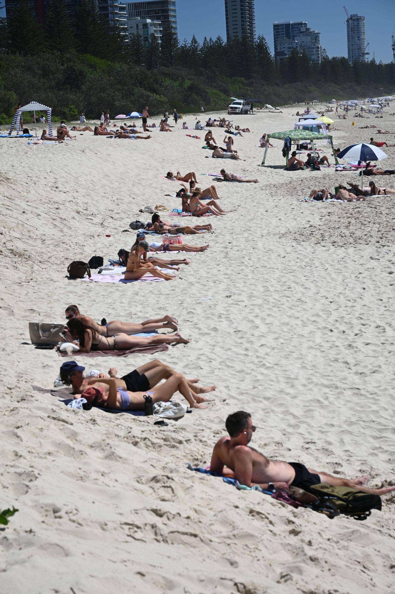 Sunbathers relaxing on a crowded sandy beach on a sunny day with city buildings visible in the background.