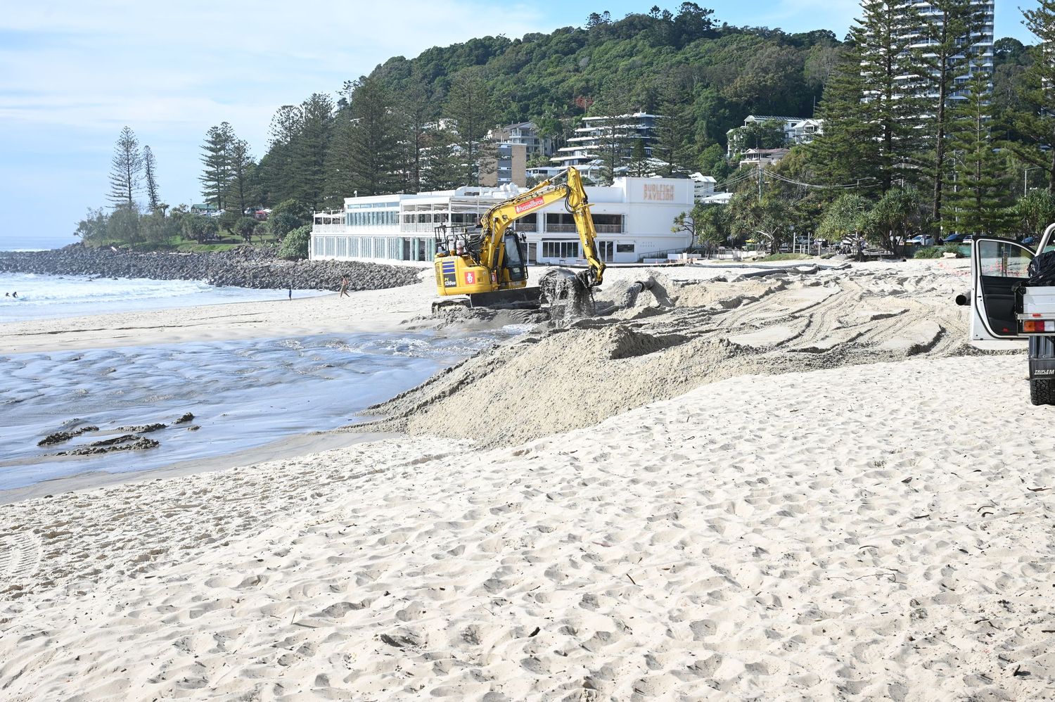 Yellow excavator reshaping sandy beach with ocean backdrop and lush green hillside in coastal town.