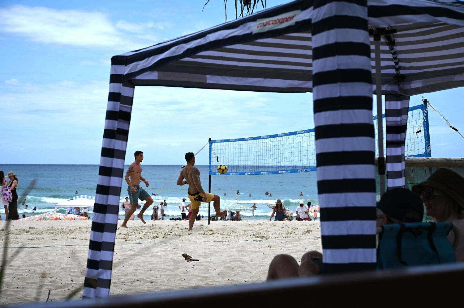People playing beach volleyball near a striped canopy with ocean views on a sunny day.