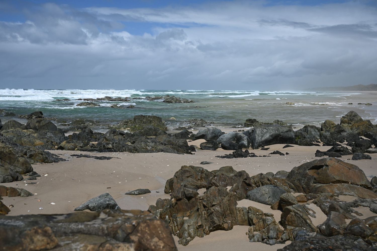 Rocky beach shoreline with crashing ocean waves under dramatic cloudy skies on a rugged coastal landscape.