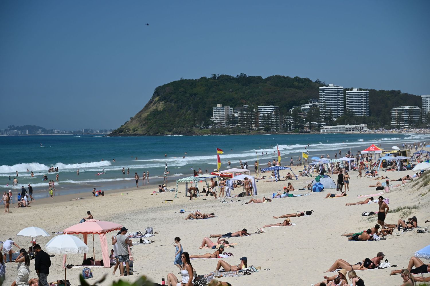 Crowded beach with sunbathers, colorful umbrellas, and calm blue ocean waves beneath a clear sky near a green headland.