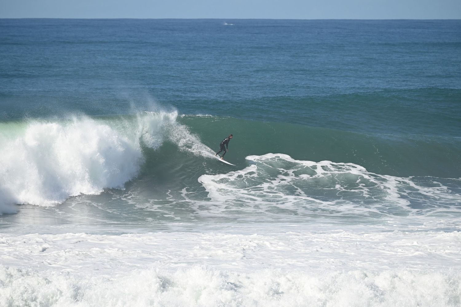 Surfer riding a massive ocean wave at a sunny beach, with white sea foam and blue water in the background.
