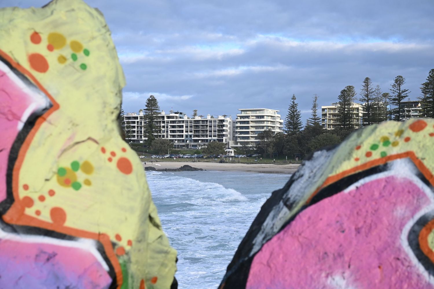 Colorful graffiti-covered rocks frame a view of beachside apartment buildings and ocean shore.