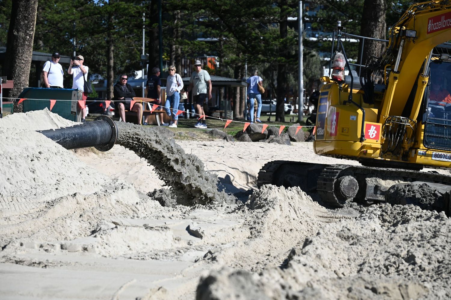 Excavator pumping sand during beach construction project with workers and trees visible in background.