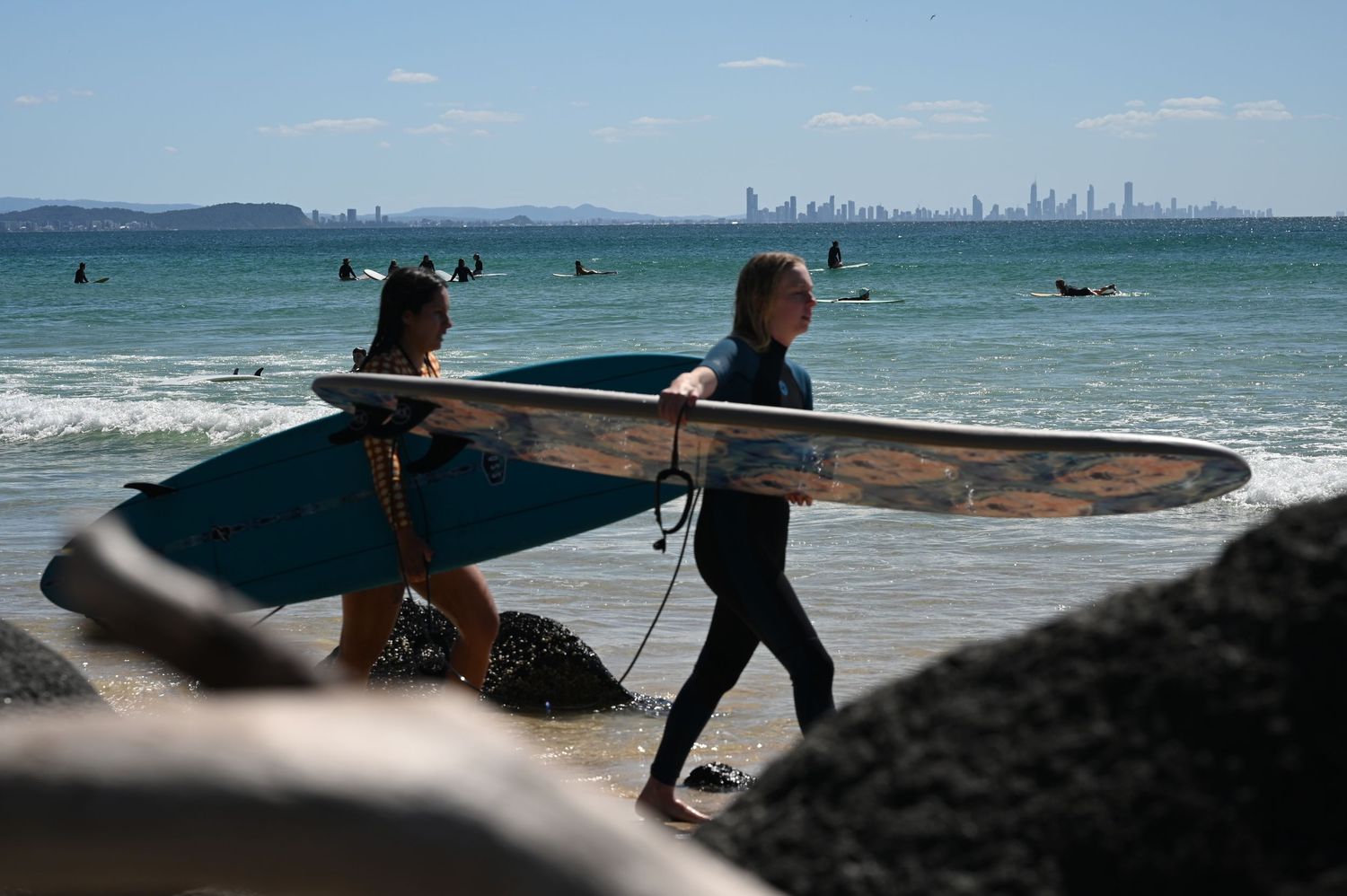 Two surfers carrying surfboards walk along a rocky beach with a city skyline visible in the background.