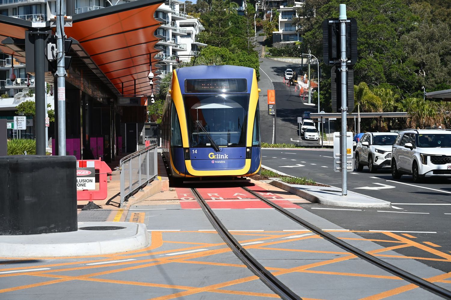 Gold Coast light rail tram arriving at a sunny street station with modern infrastructure and parked cars nearby.