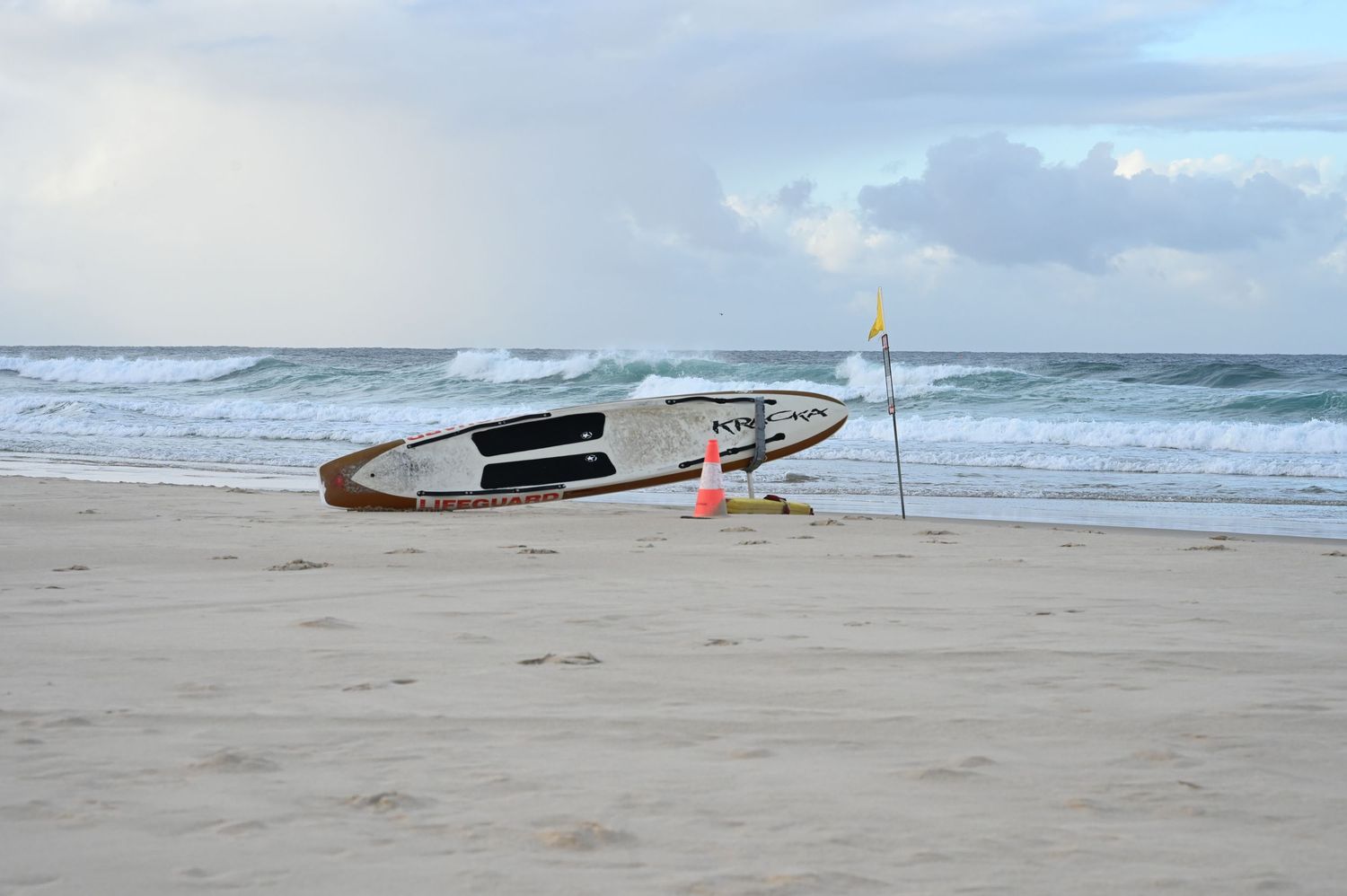A surfboard resting on sandy beach near orange cones and a flag, with ocean waves in the background on a cloudy day.