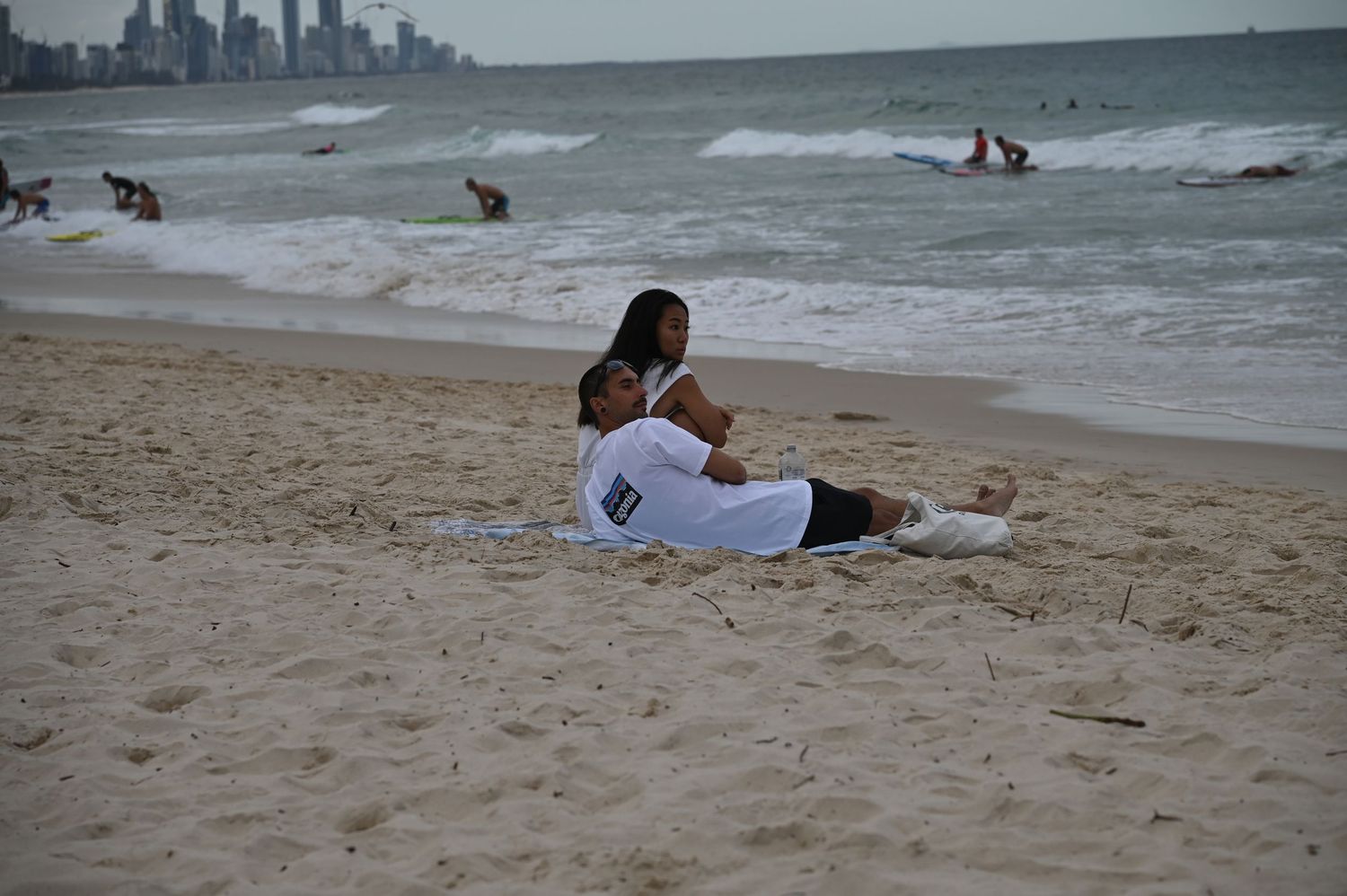Couple relaxing together on sandy beach with ocean waves and city skyline visible in background.
