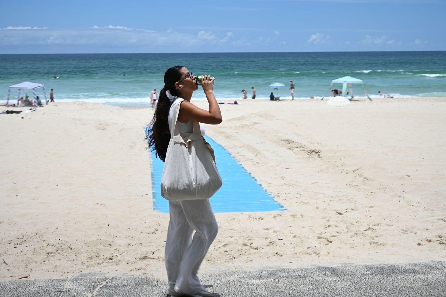 Woman in white outfit drinking from a bottle on a sunny beach with blue ocean waves in the background.