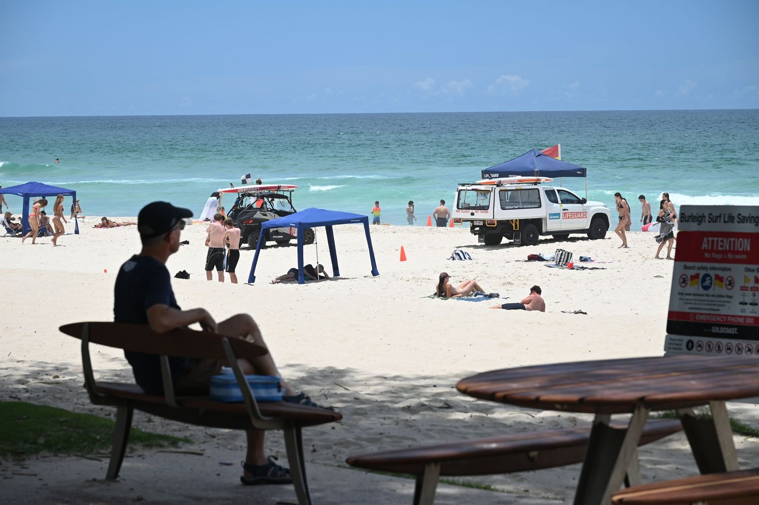 A man sits on a wooden bench overlooking a sunny beach with lifeguard vehicles, sunbathers, and ocean waves in the background.