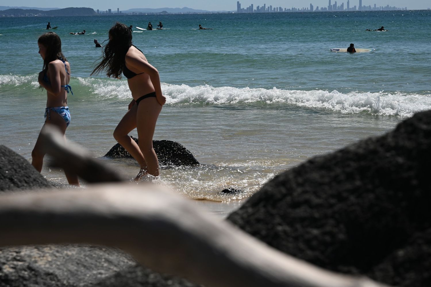 Two women in bikinis walk along a rocky beach with surfers and a city skyline visible in the background.