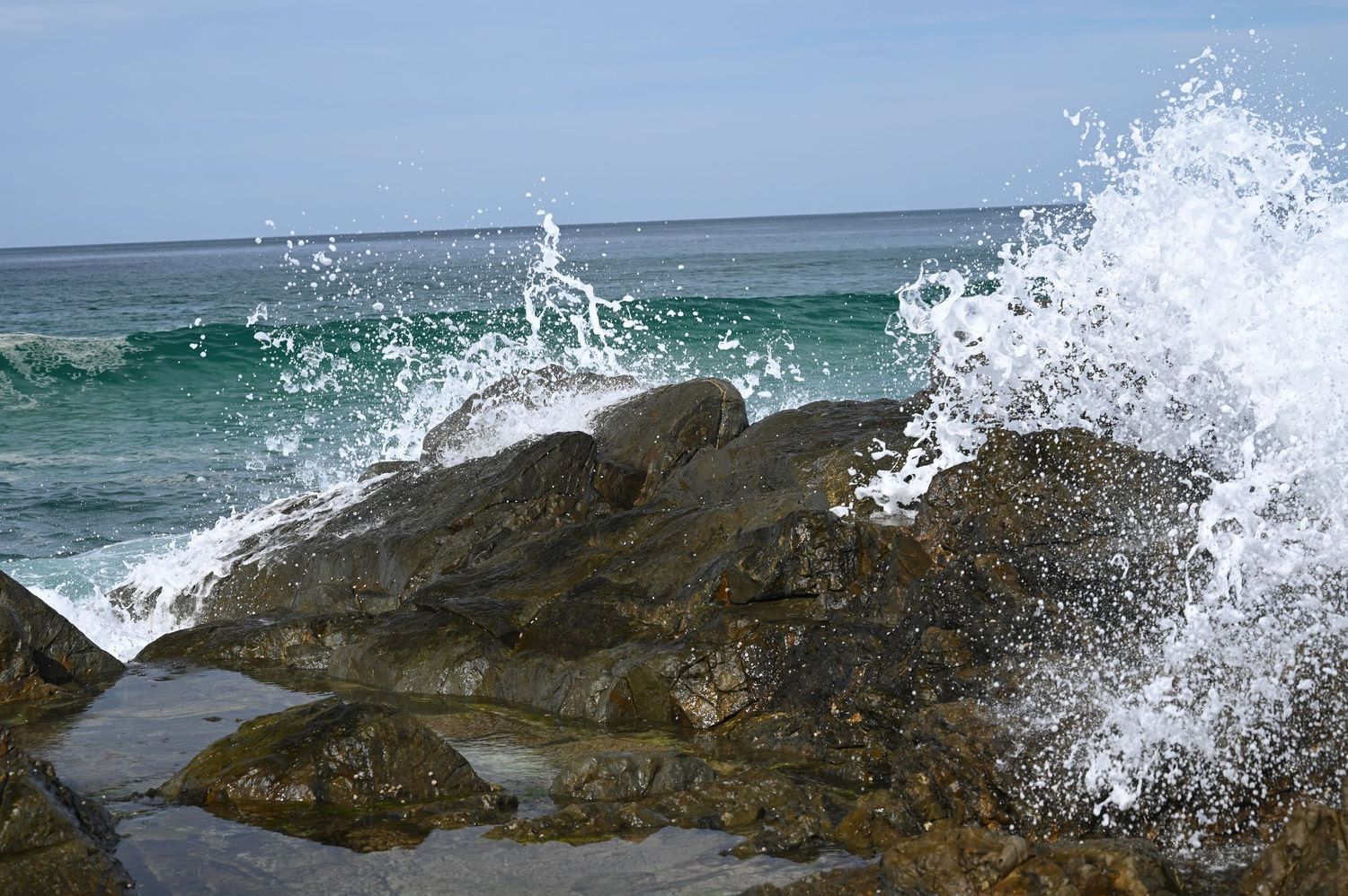 Ocean waves crashing dramatically against rocky shore with white sea spray, blue water and clear sky in background.