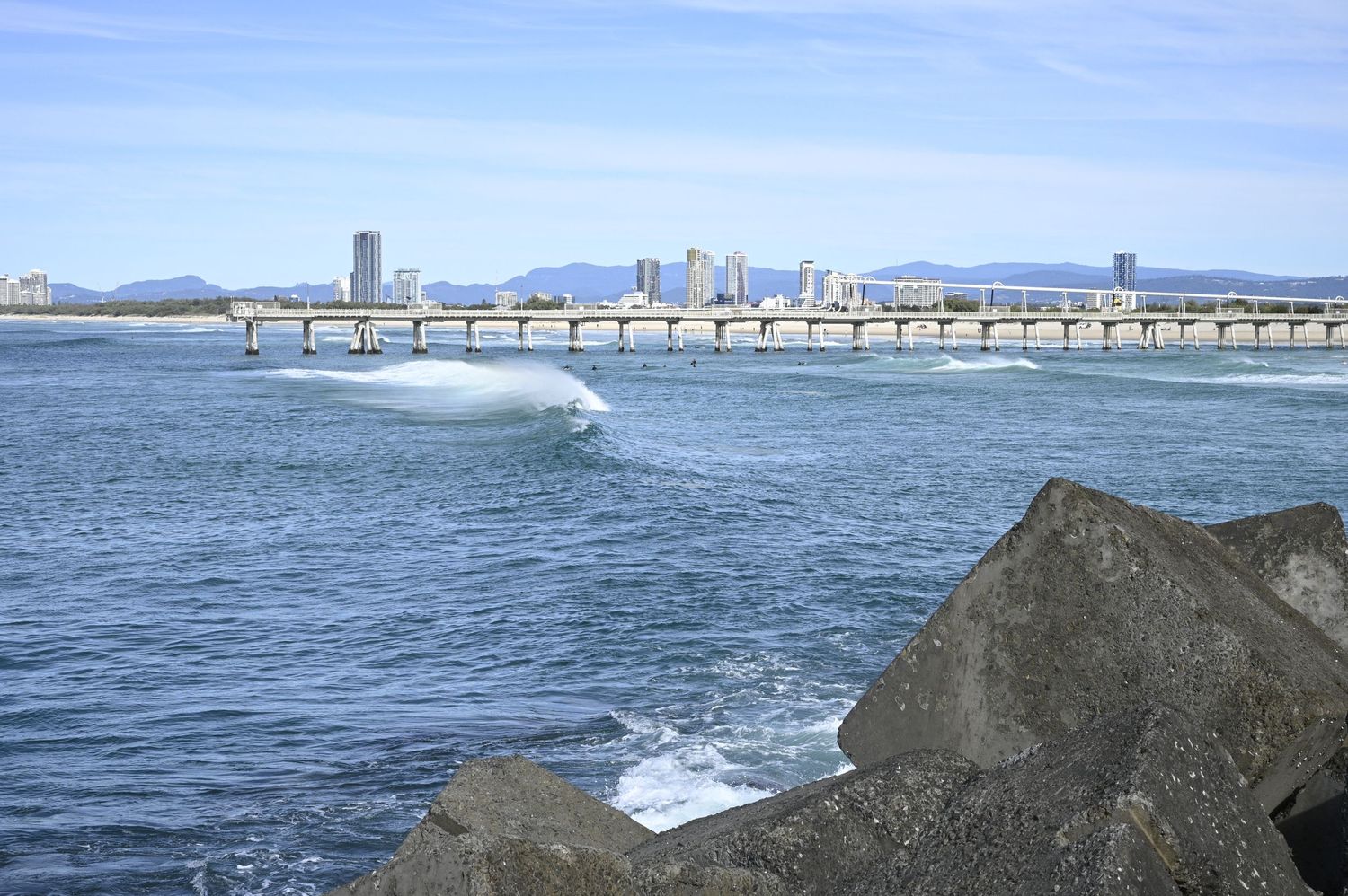 Surfer riding a wave near a bridge with a Gold Coast skyline and mountains in the background on a clear day.