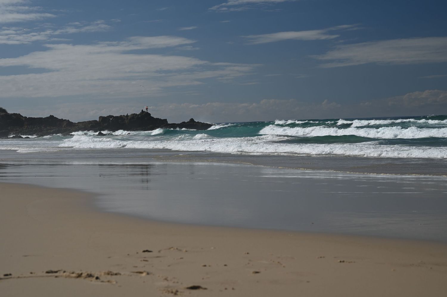 Sandy beach with rolling white waves crashing on shore, rocky outcrops visible, and a partly cloudy blue sky above the ocean.