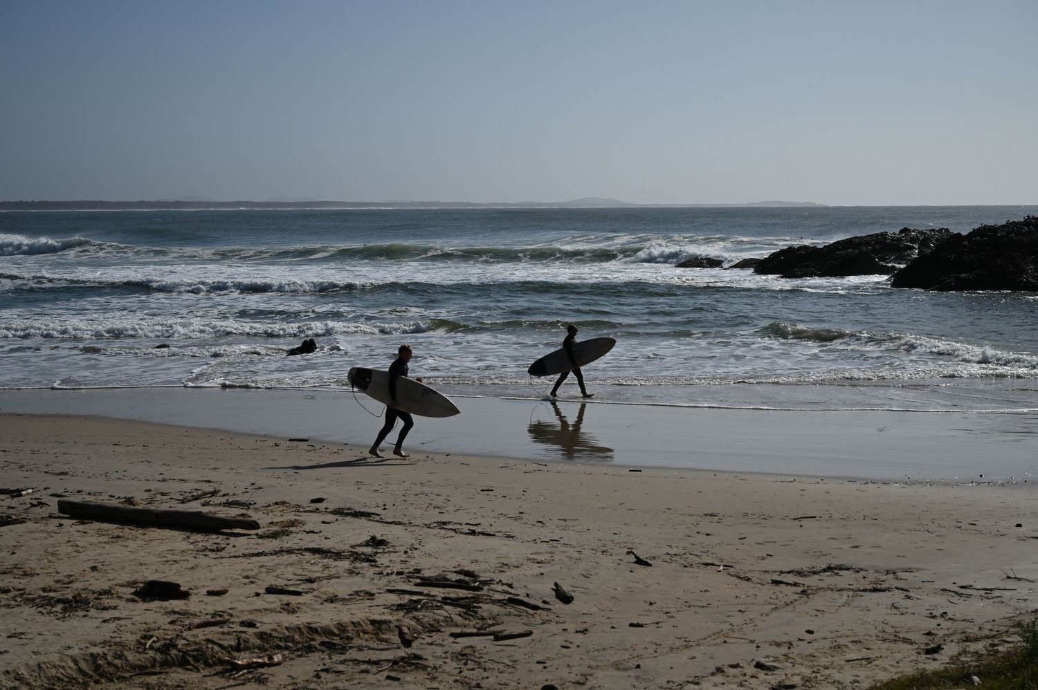 Two surfers carrying boards walk along a sandy beach toward ocean waves under a clear blue sky.