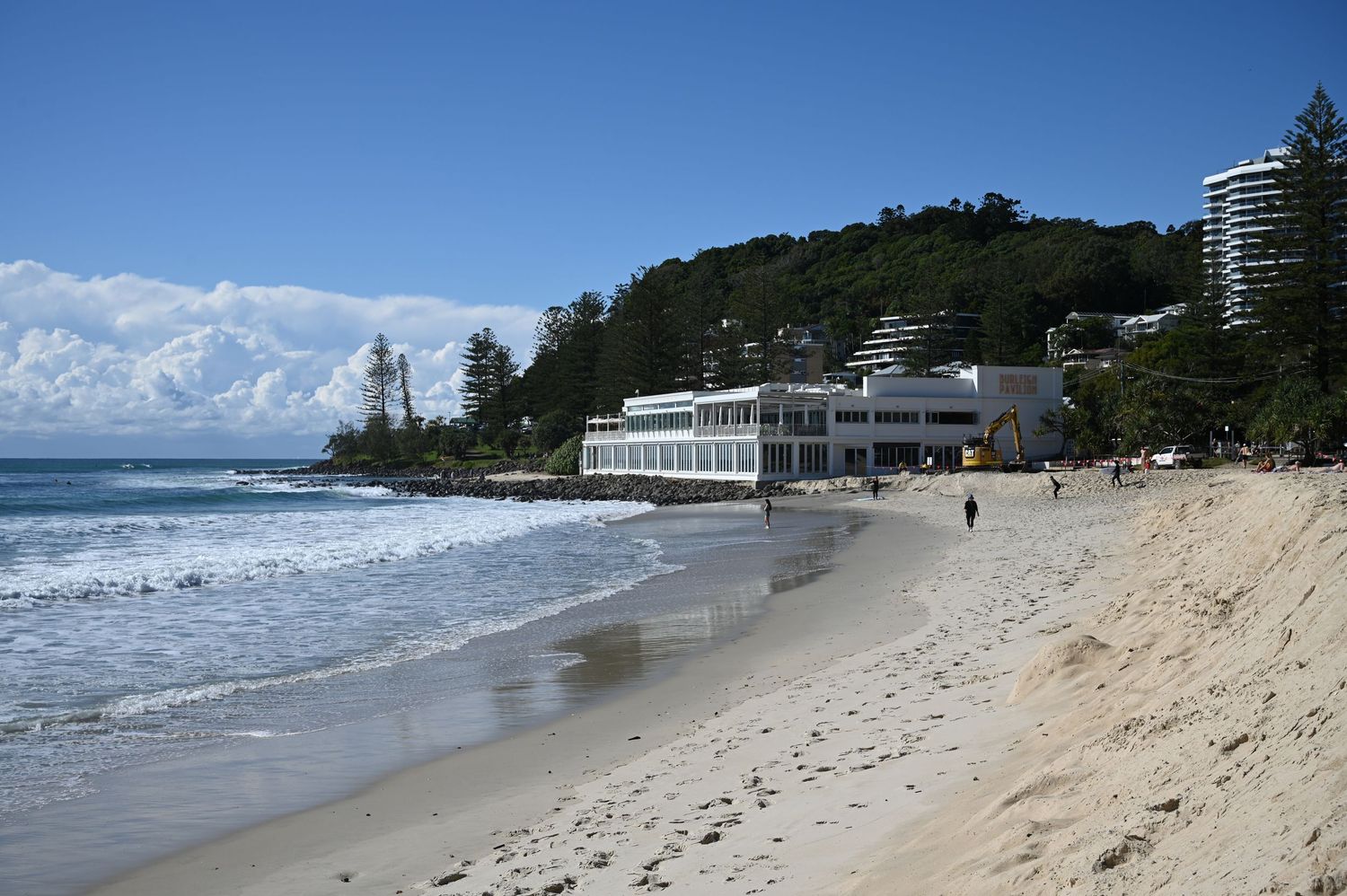 Sandy beach with white building and tree-covered headland under blue sky in Burleigh Australia.