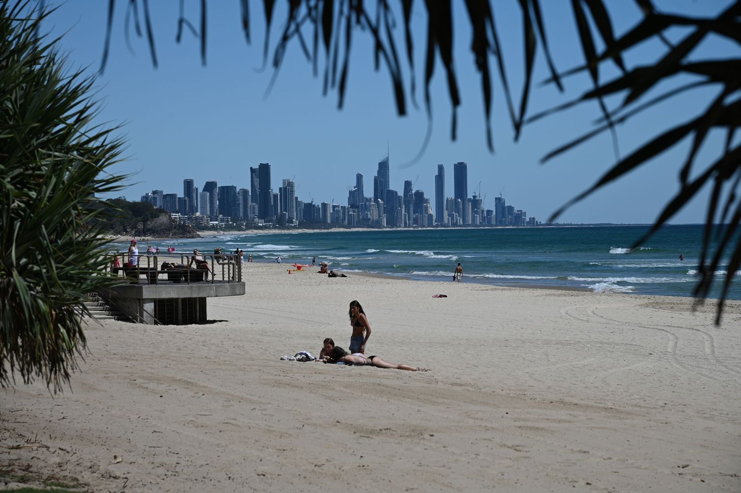 Sunbathers relax on a sandy beach with a stunning city skyline visible across the turquoise ocean on a clear sunny day.