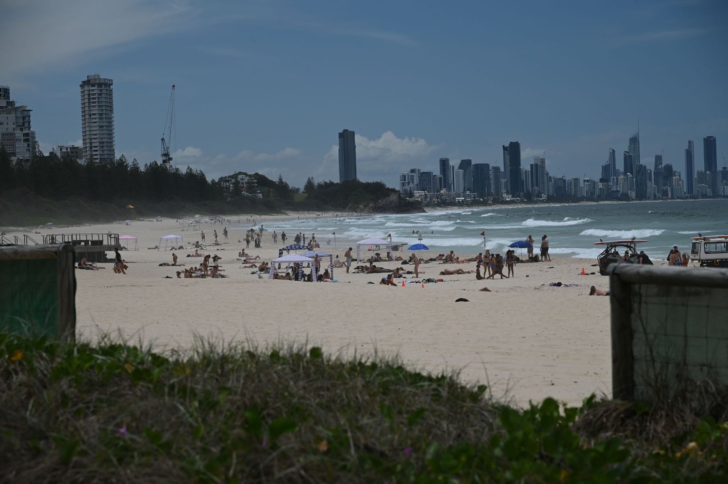 Busy beach scene with white sand, sunbathers, and a stunning city skyline backdrop under a partly cloudy sky.