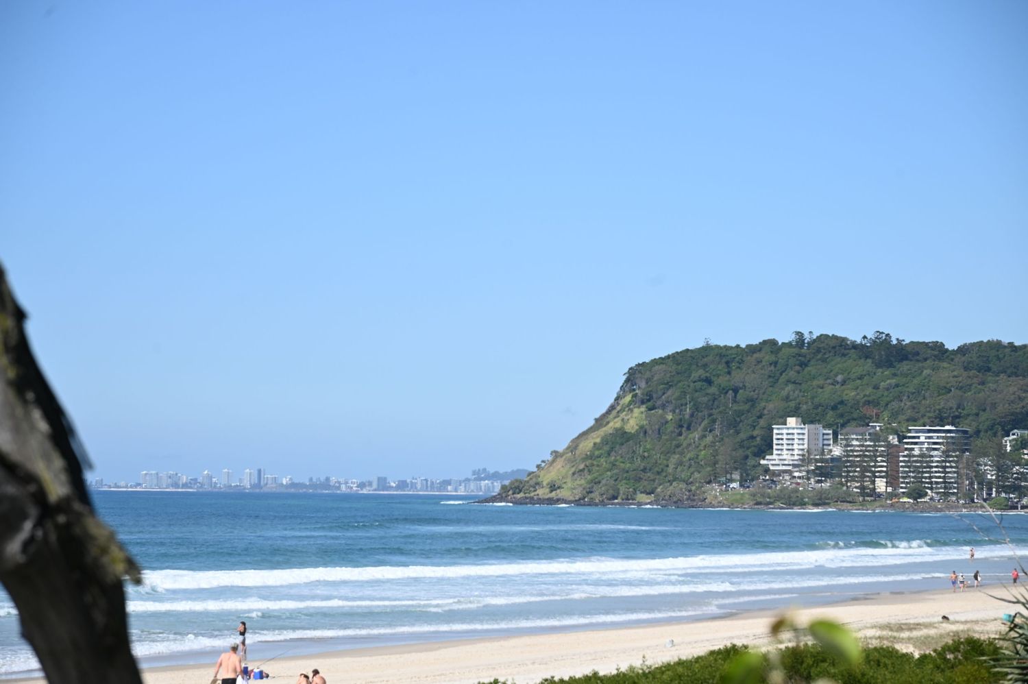Scenic beach with golden sand, blue ocean waves, and a lush green headland under a clear sky.