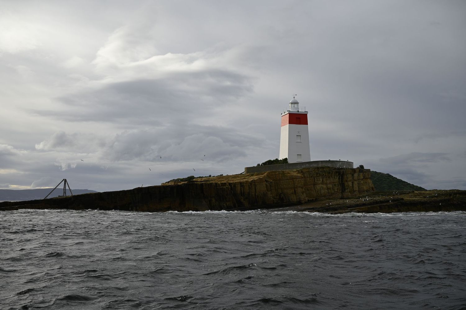 A red and white lighthouse perched on a rocky island surrounded by choppy ocean waters.