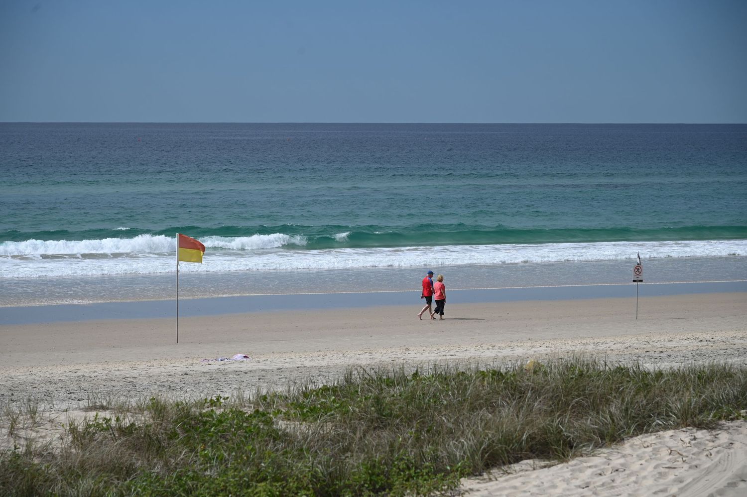 Two lifeguards walk on a sandy beach with yellow-red flag, turquoise waves, and clear blue sky.