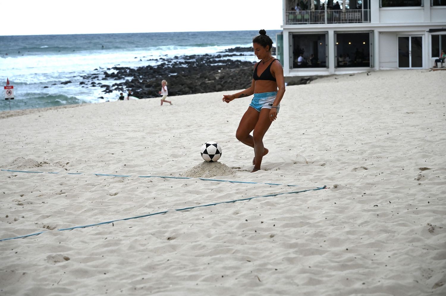 Woman in bikini playing foot volleyball on sandy beach with ocean waves in the background.