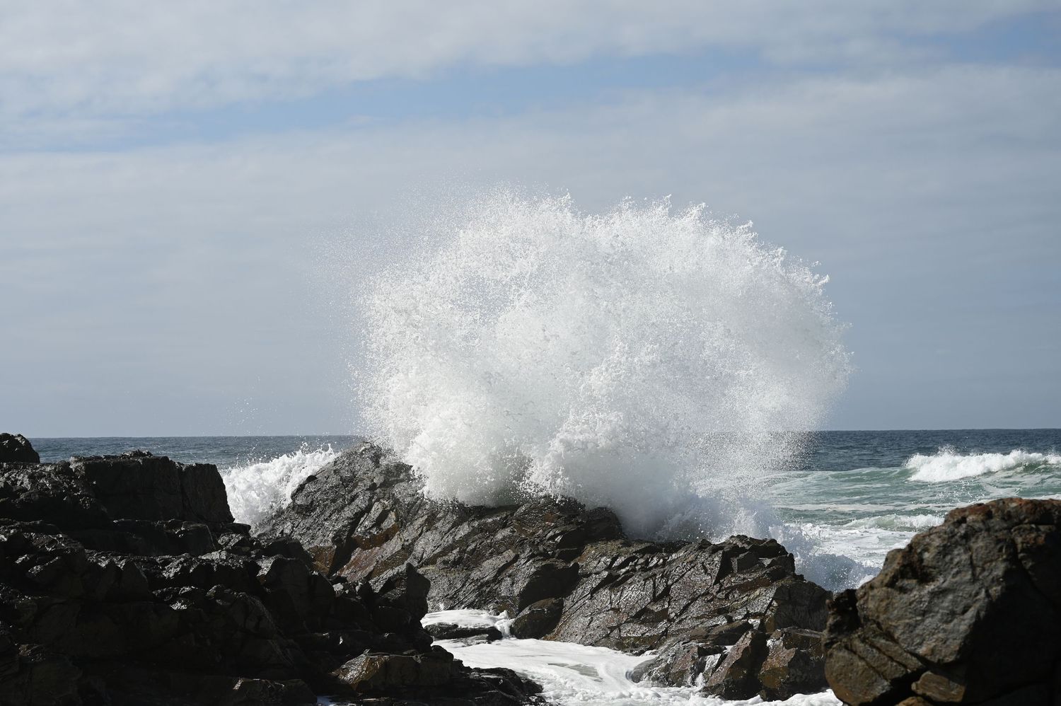 Ocean waves crashing dramatically against rocky shoreline, sending white sea spray high into the air under a cloudy sky.