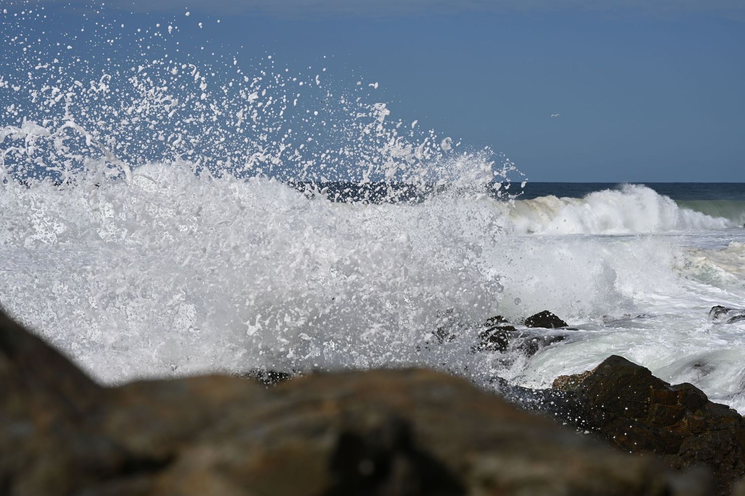 Powerful ocean waves crashing against rocky shoreline with white sea spray under a clear blue sky.