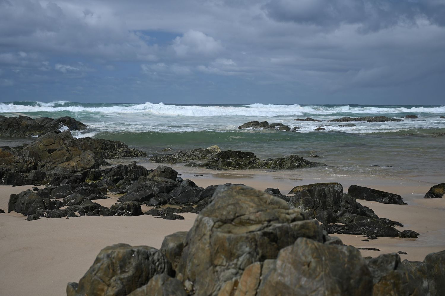 Rocky beach with crashing waves under stormy skies, featuring dark boulders on sandy shore with turbulent ocean waters.