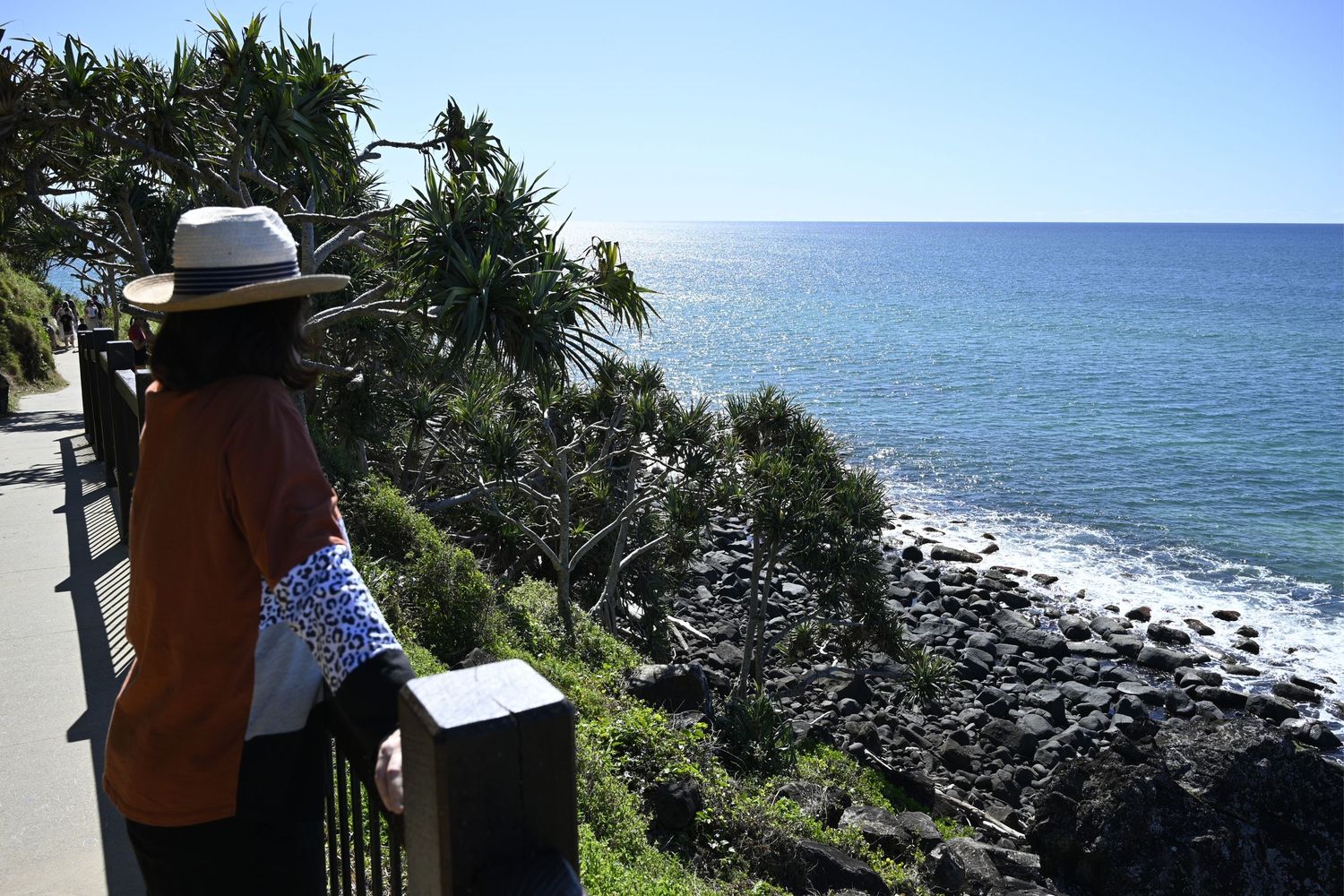 Woman in hat gazing at rocky coastline with turquoise ocean views from scenic coastal walkway.