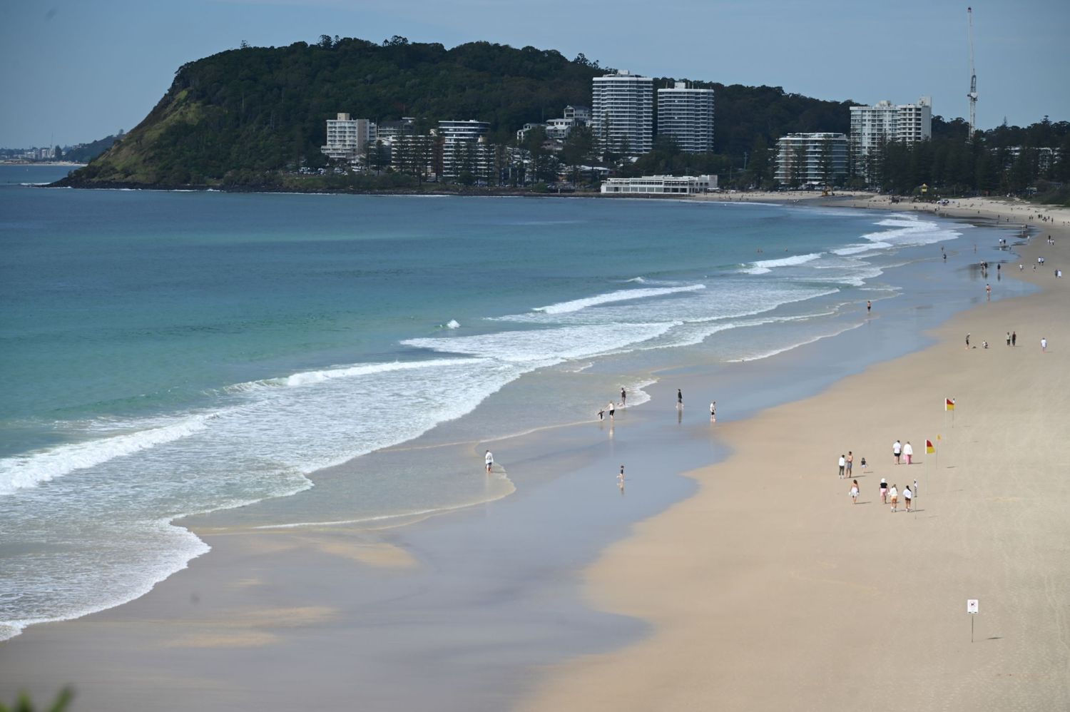 Aerial view of a long sandy beach with gentle waves, scattered swimmers, and a green headland with buildings in the background.