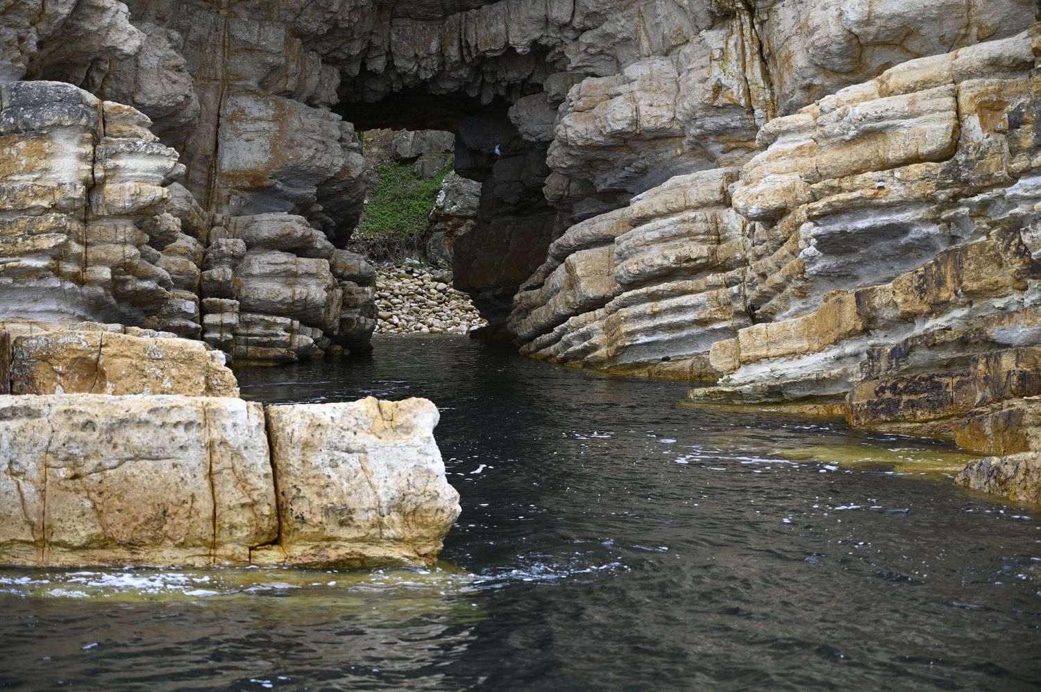 Rocky coastal cave entrance with layered limestone cliffs and dark tidal waters flowing through a narrow sea cave opening.
