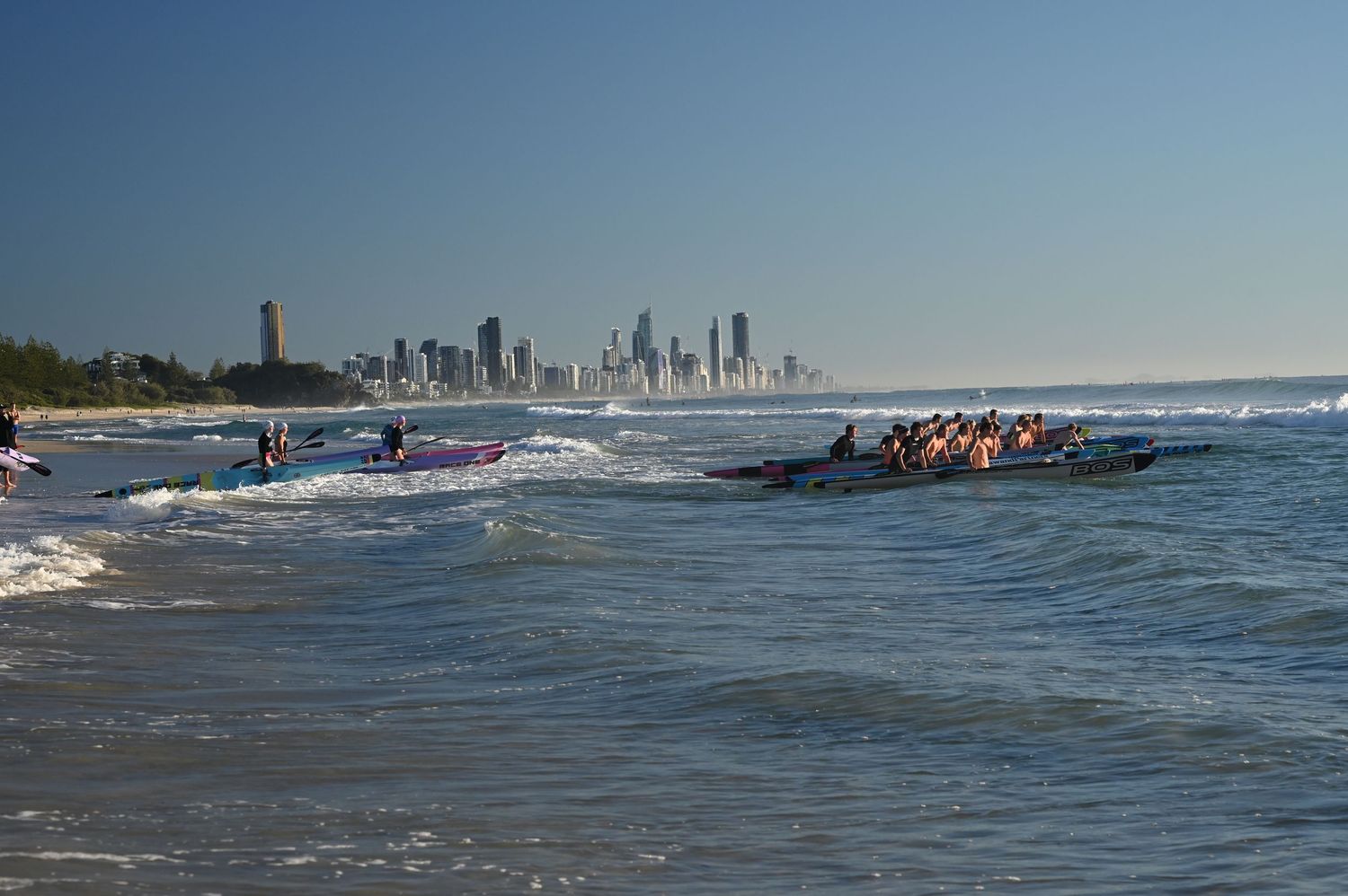 Surf boats racing through ocean waves near Gold Coast skyline on a clear sunny day.