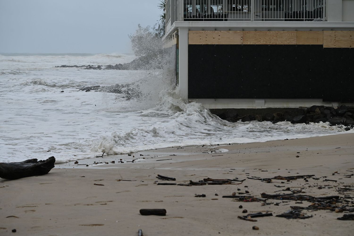 Rough storm waves crashing against a beachfront building with debris scattered on the sandy shore.
