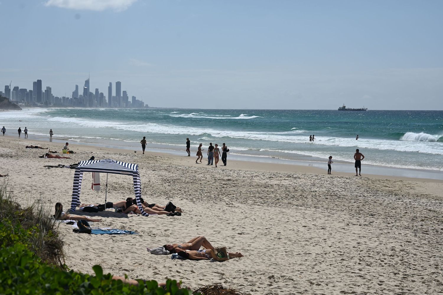Sunny beach scene with sunbathers, a striped cabana, swimmers, and a city skyline visible in the background.