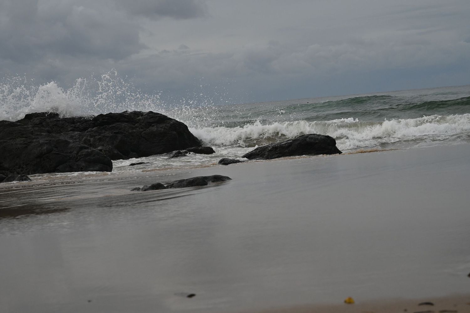 Rocky shoreline with crashing ocean waves on a cloudy day, with dark boulders and white sea foam on a sandy beach.