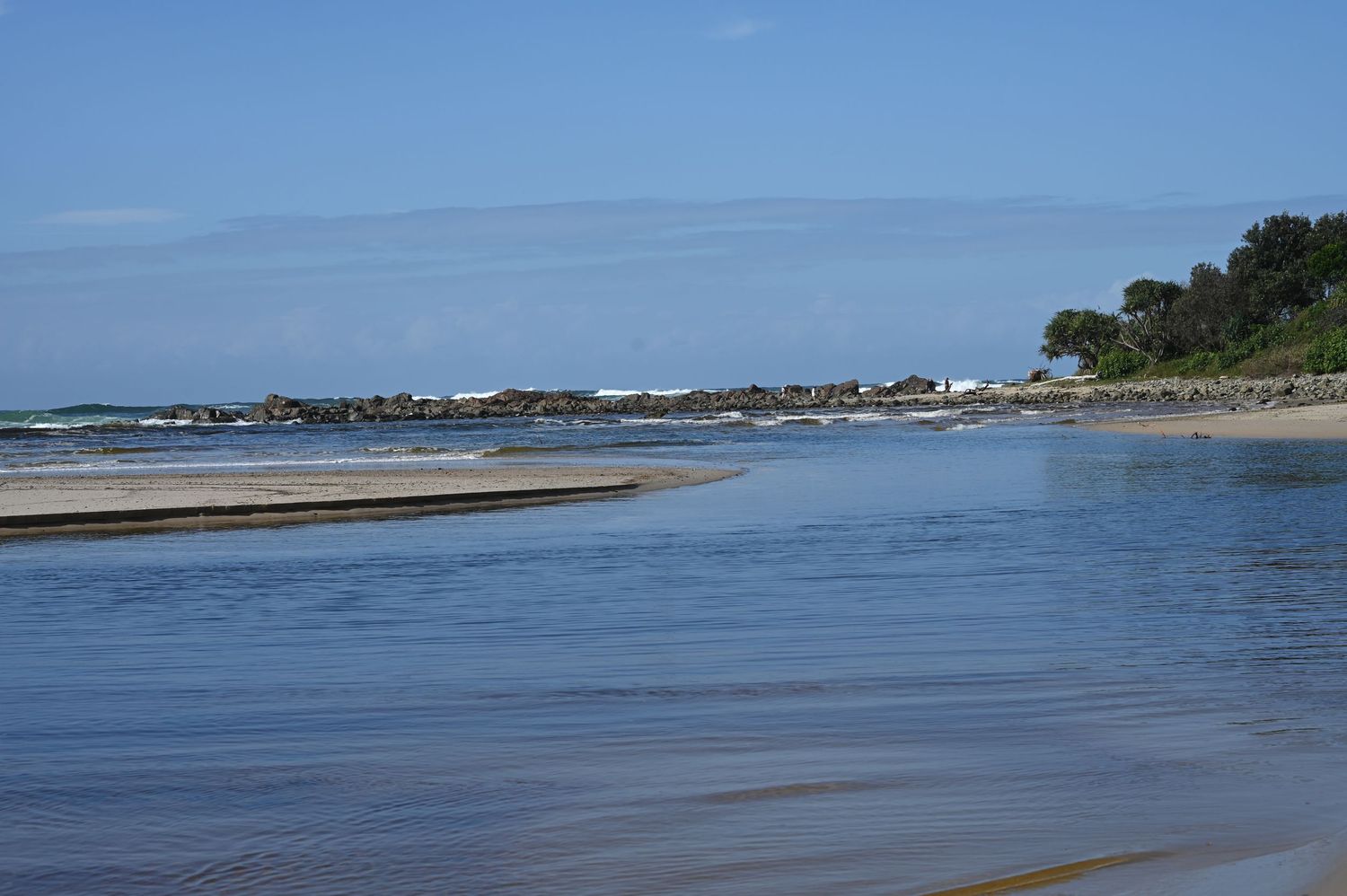 Calm coastal beach scene with shallow tidal waters, rocky shoreline, and lush green trees under a clear blue sky.