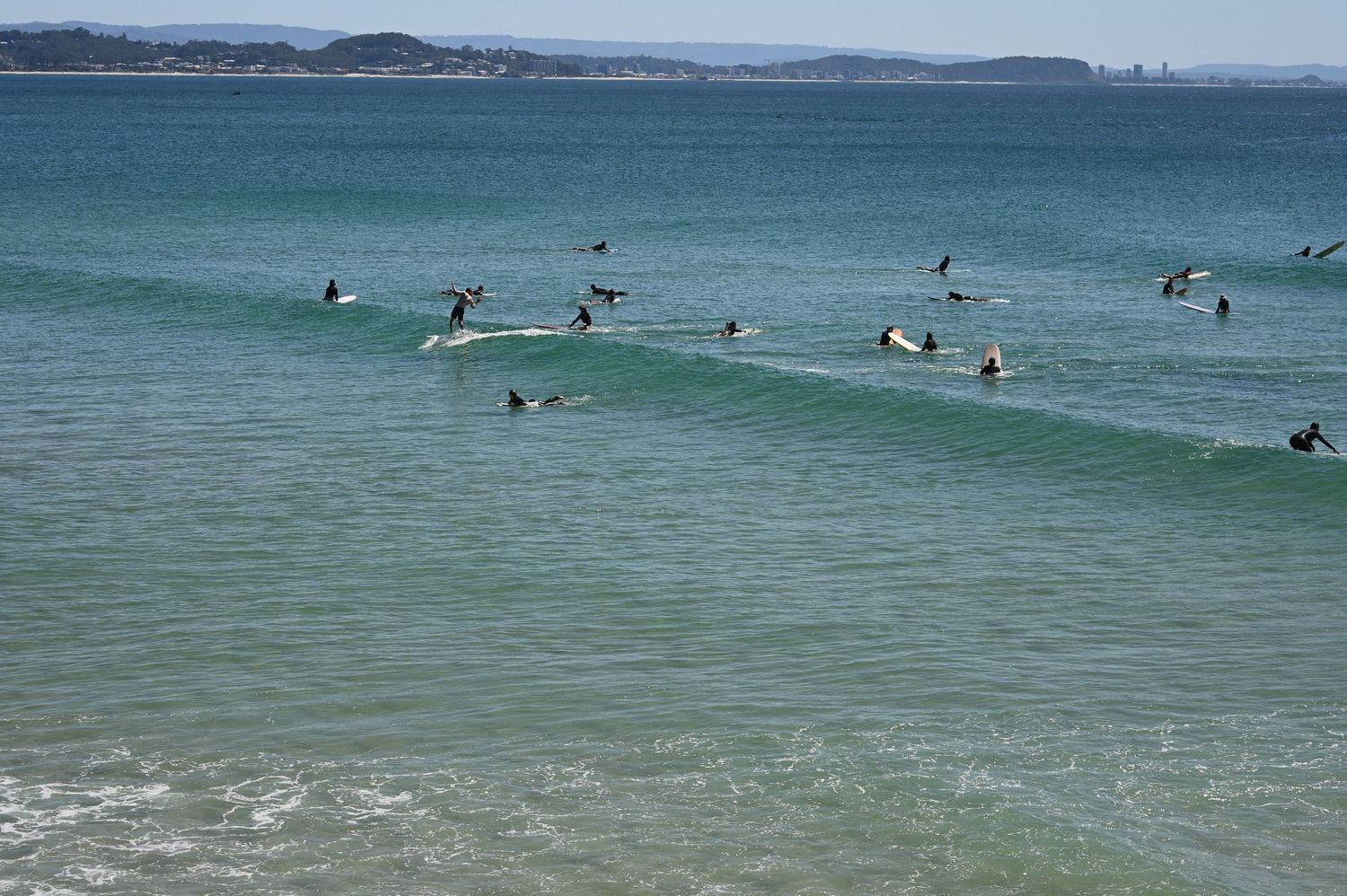 Group of surfers paddling and riding waves on a clear turquoise ocean near a sandy beach on a sunny day.