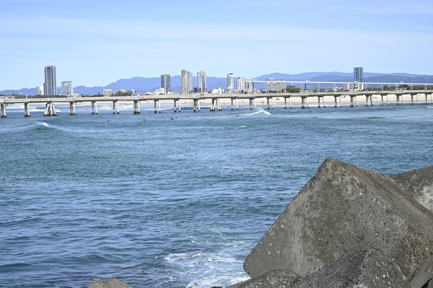 Coastal city skyline with a long bridge over blue ocean waters, rocky foreground, and mountains in the background.