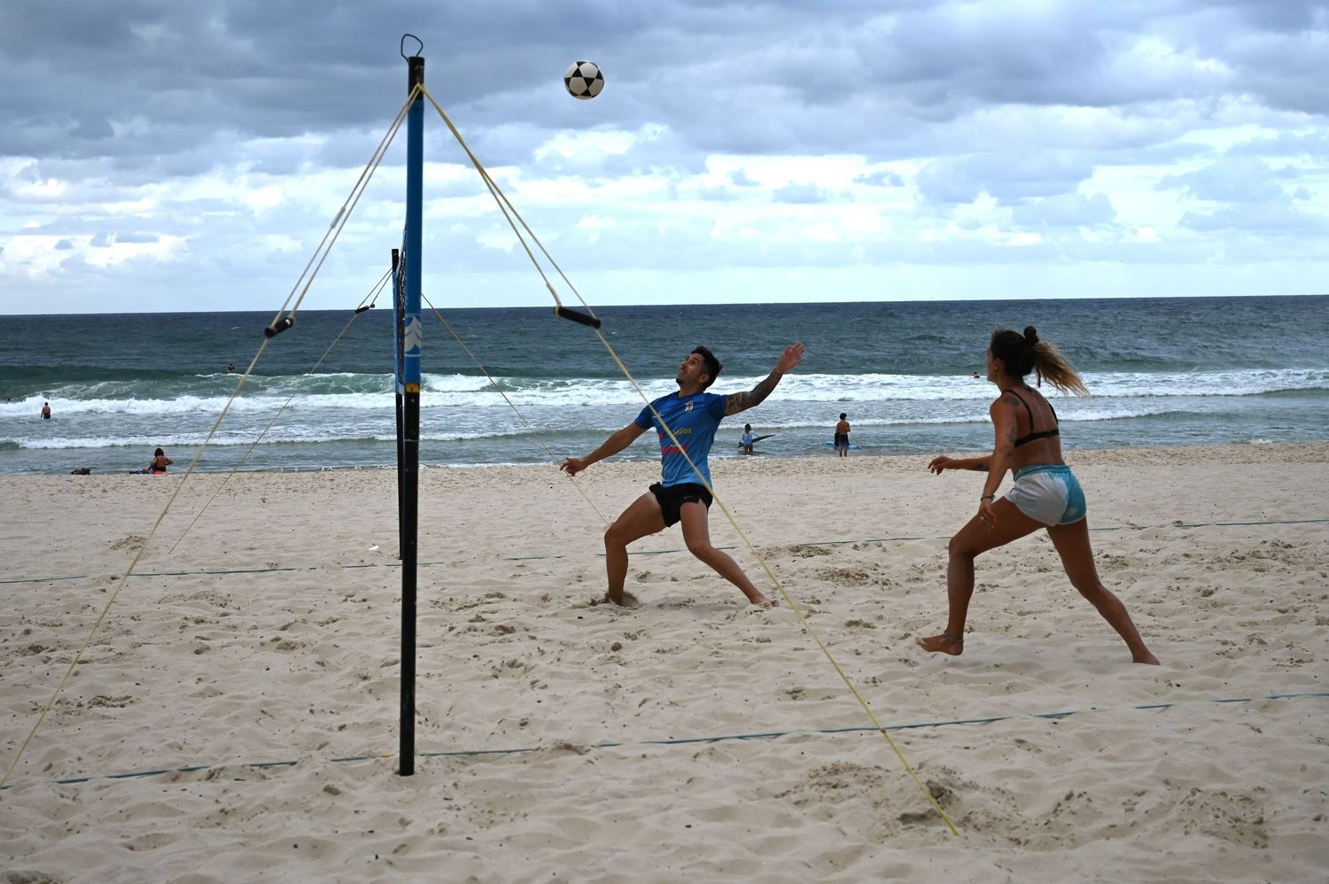 Two players enjoy a beach volleyball game on a sandy shore with ocean waves and cloudy skies in the background.